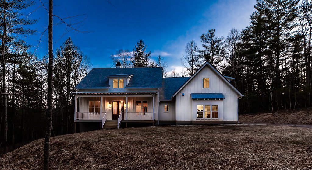 White farmhouse at dusk with lit windows and a metal roof among tall pines