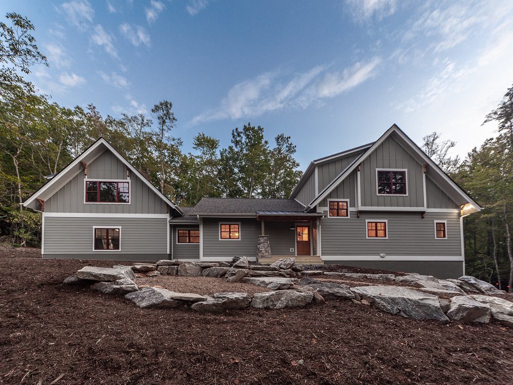 Gray board-and-batten home at dusk with stone landscaping and warmly lit windows