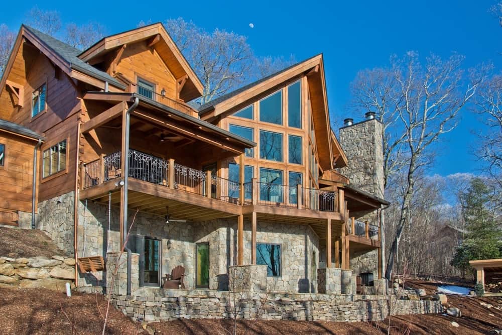 Side view of the timber home with tall windows, a stone foundation and a covered deck