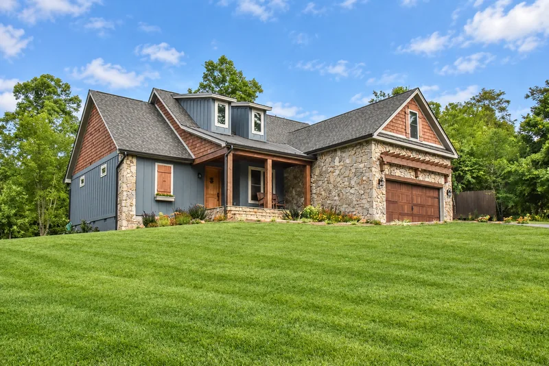 Blue-gray and stone craftsman home with wooden front door and garage doors on a green sloping lawn
